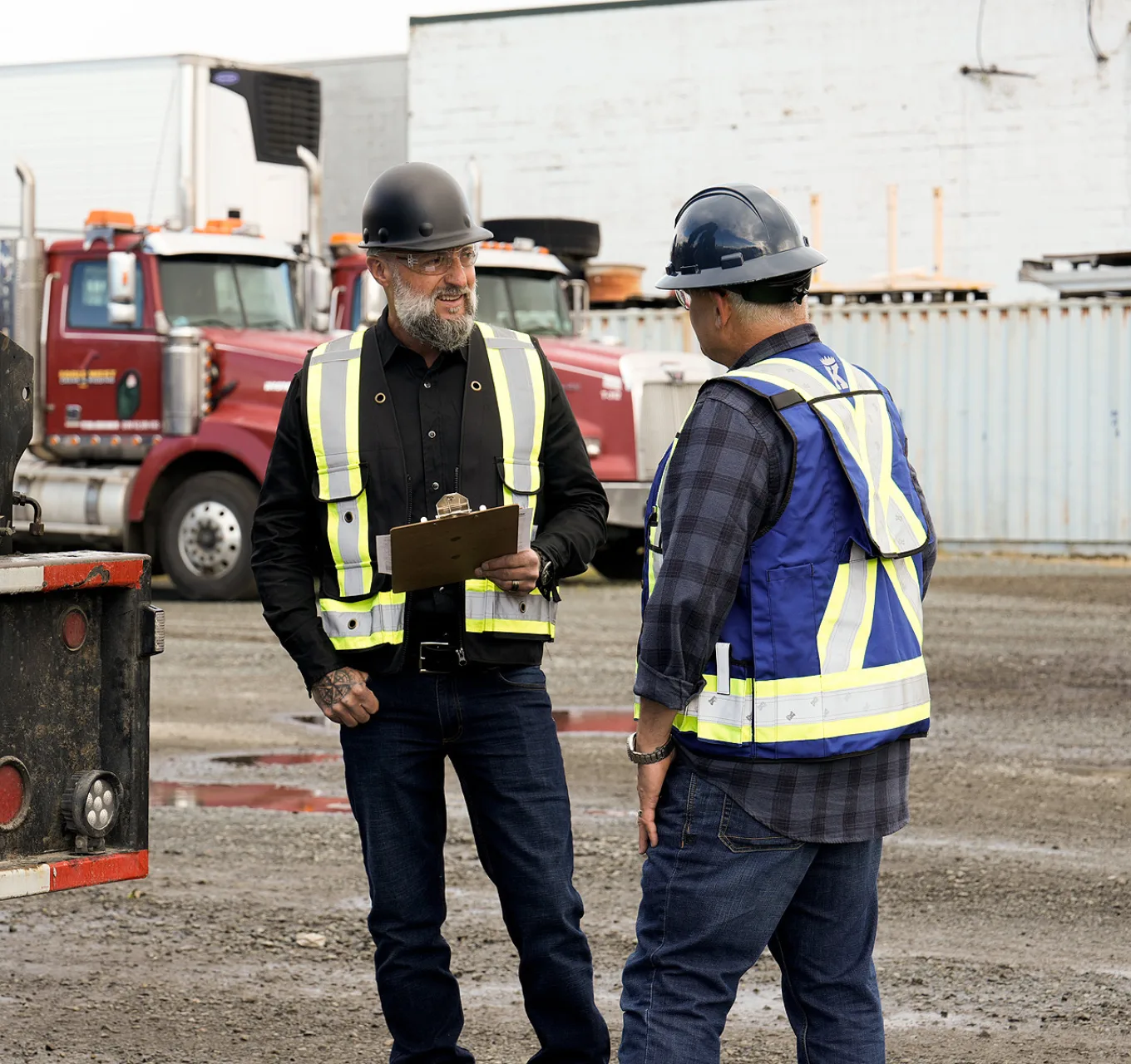 An employer holding a clipboard talka with a worker outdoors near a crane.