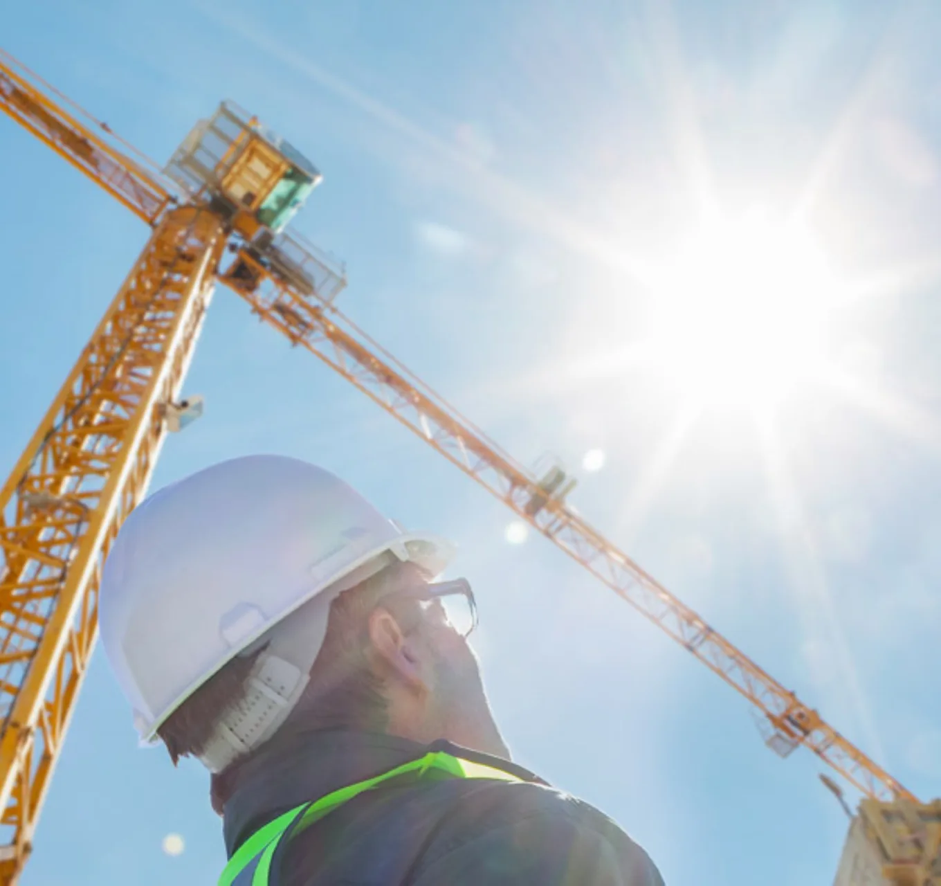 A worker looks up at sun appearing next to a tower crane on a bright day.