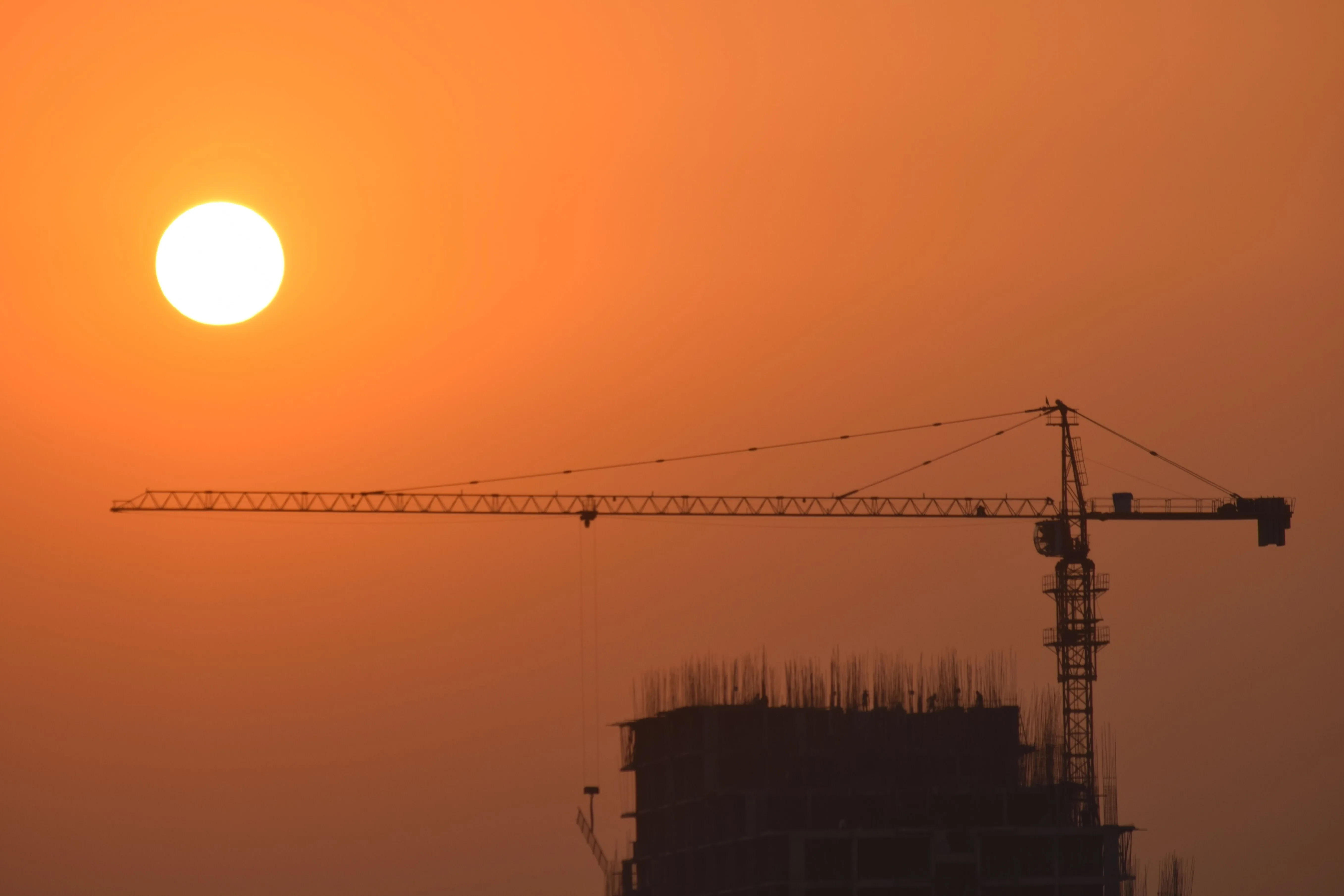 An orange sky with the sun and the silhouette of a tower crane and an unfinished building.