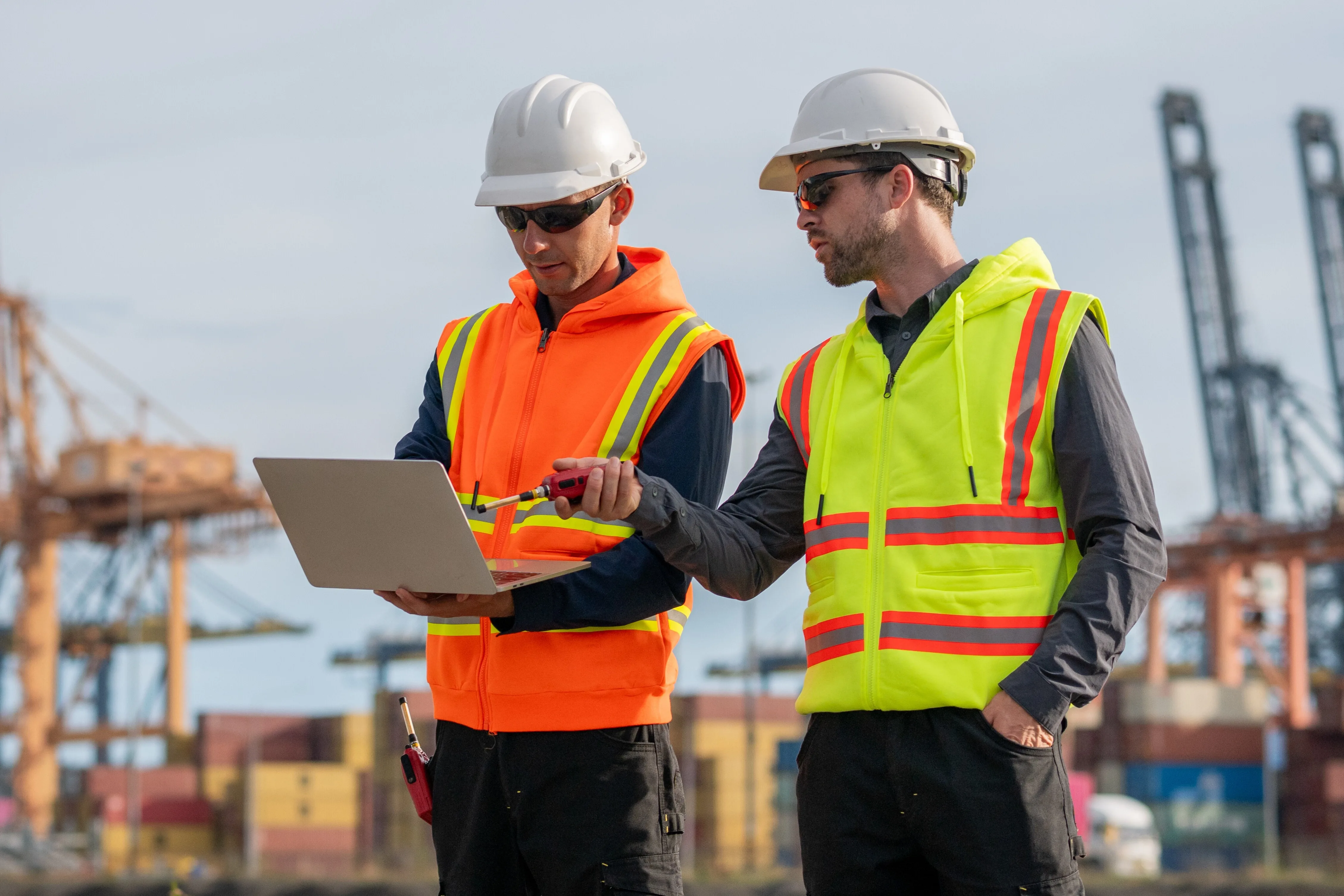 Two male workers wearing personal protective equipment look over the information on a laptop.