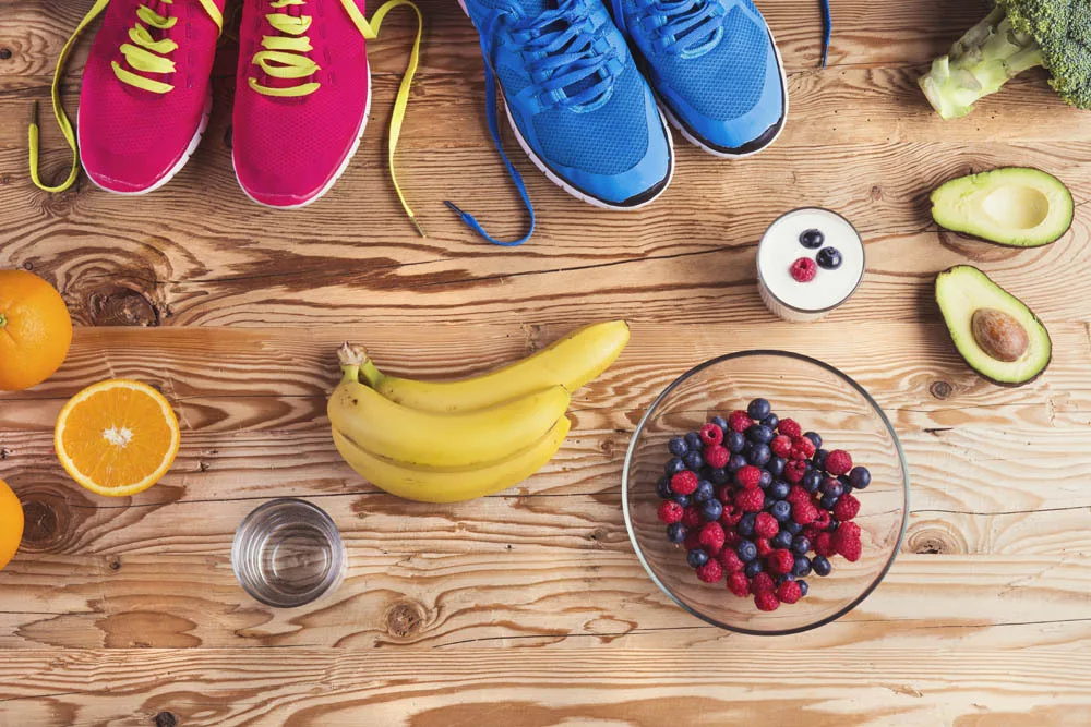 A table with two pairs of brightly coloured excercise runners and a variety of healthy foods including oranges, bananas, mixed berries, avacado and broccoli.