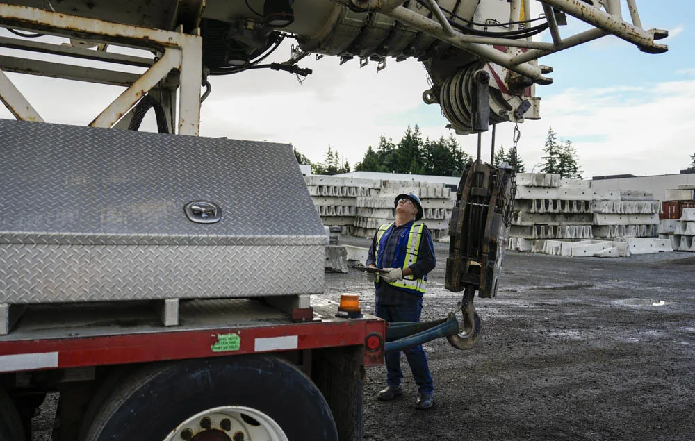A crane operator wearing personal protective equipment and holding a clipboard looks overhead to inspect the boom of a large mobile crane.