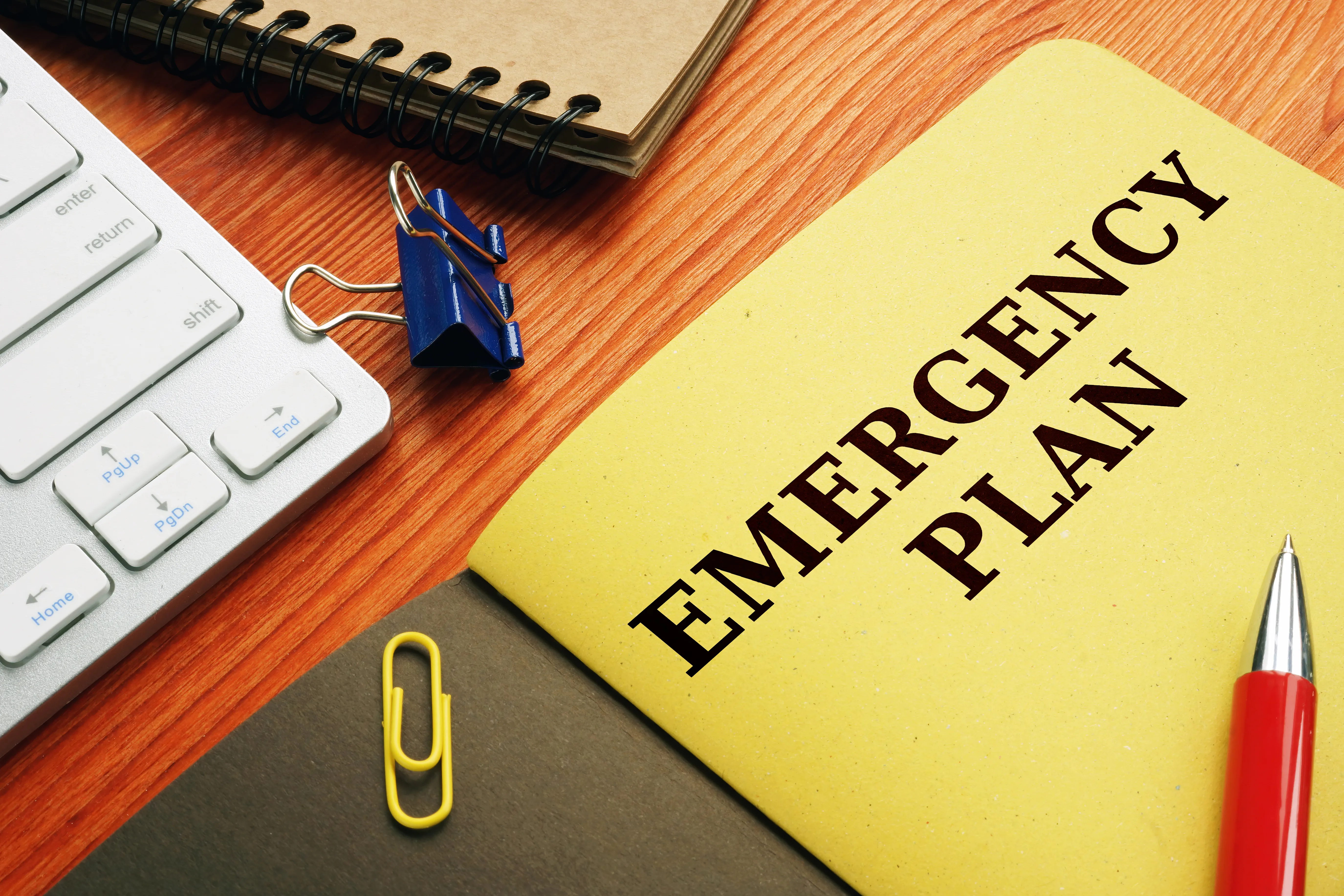 A yellow emergency plan folder on a desk with a red pen, paper clips and part of a keyboard showing.