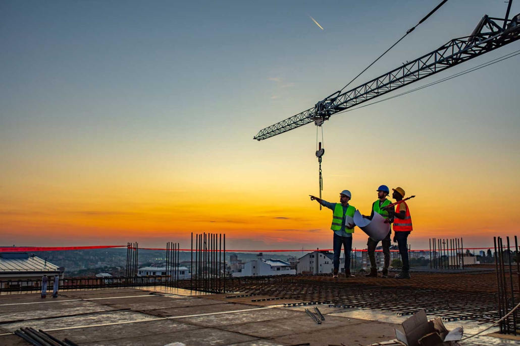 3 construction workers on top of a building being constructed with a crane in the background at sunset.