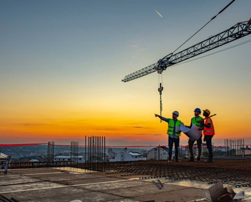 3 construction workers on top of a building being constructed with a crane in the background at sunset.