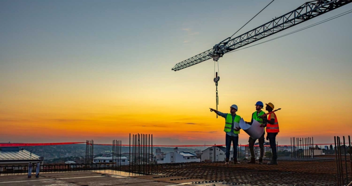 3 construction workers on top of a building being constructed with a crane in the background at sunset.