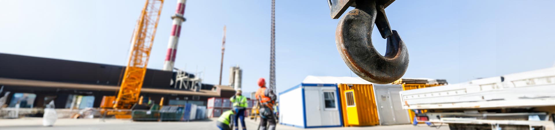 3 men fully dressed in PPC on a construction site look upwards and are out of focus. A crane hook nearer to the camera is in focus.